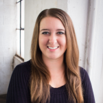 woman wearing black standing in front of a white wall. She has long brown hair